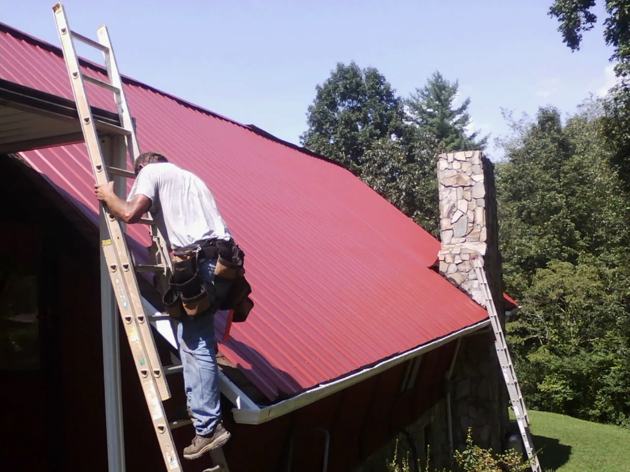 Roofer on a ladder beside a red metal roof and stone chimney — Soffit & Fascia Repair by Roofing Brothers in West Milford